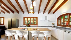 Cozinha moderna com vigas de madeira, armários brancos, uma ilha central com banquetas e uma janela grande com vista para o verde.