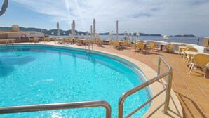 Ein Pool mit klarem, blauem Wasser, umgeben von gelben Liegestühlen und Sonnenschirmen, mit Blick auf das Meer und Hügel im Hintergrund.