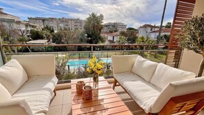 Ein Balkon mit zwei weißen, gepolsterten Sofas, einem Holztisch und einer Vase mit gelben Blumen. Im Hintergrund ist ein Schwimmbecken zu sehen.