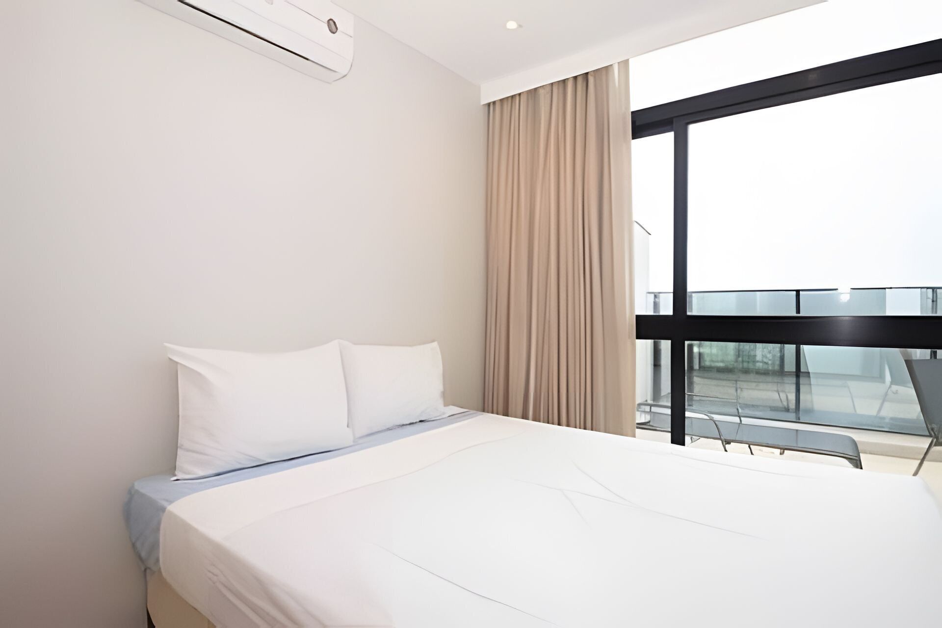 A modern hotel room with a large bed, a window with a view of a building, and a balcony with glass railings.