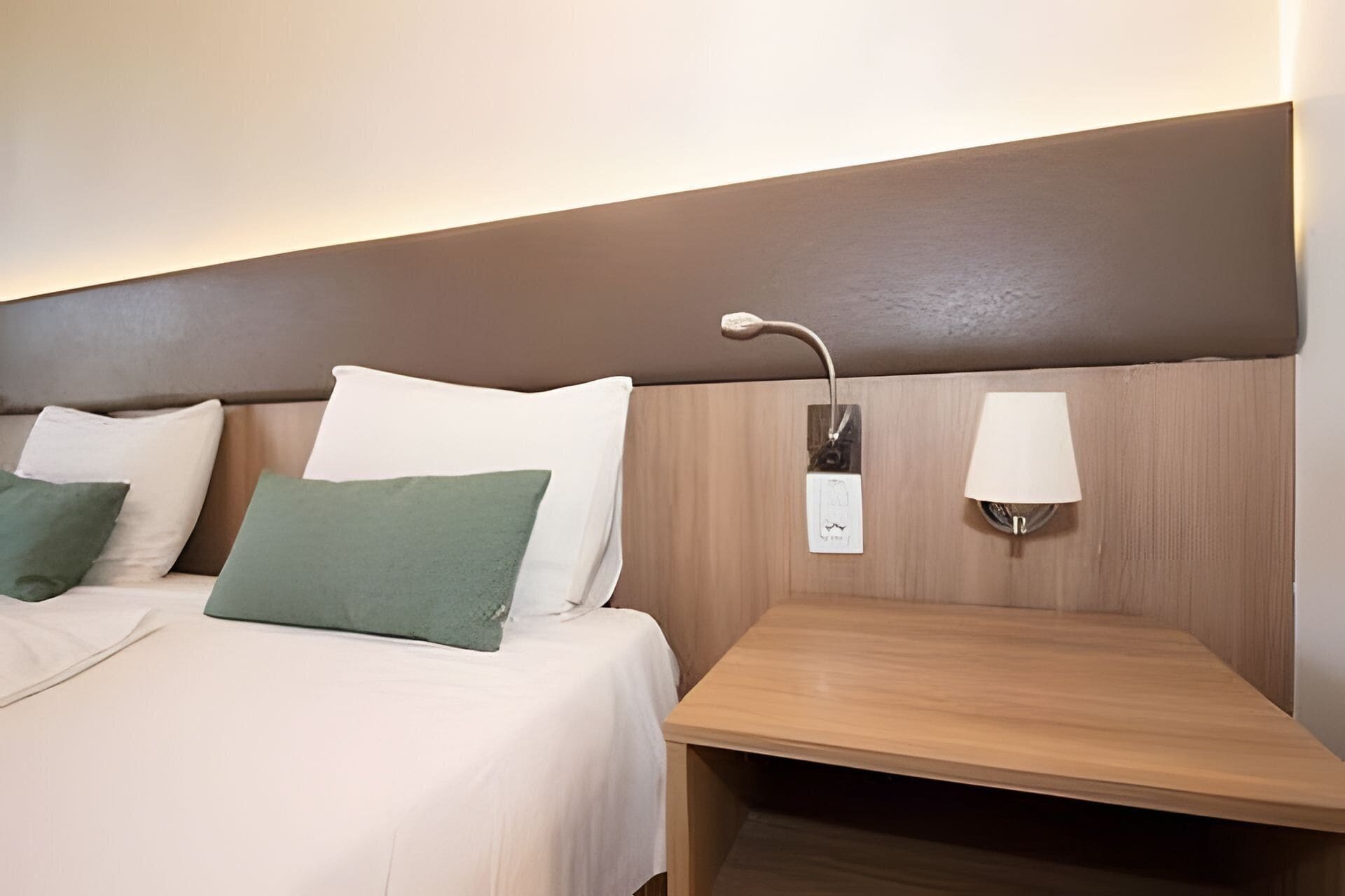 Una habitación de hotel con una cama, almohadas, una mesita de noche y una lámpara de pared.