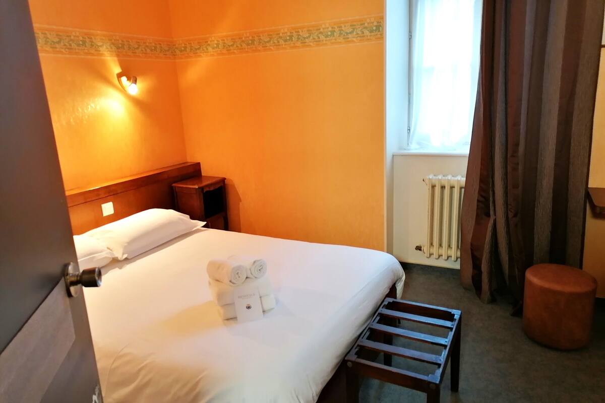 A hotel room with a bed, a nightstand, a radiator, and a window with curtains.