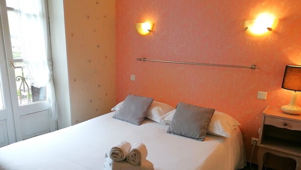 A neatly made bed with pillows, a bedside table with a lamp, and a window with curtains. - Hôtel Aux Armes d'Estaing (Estaing)