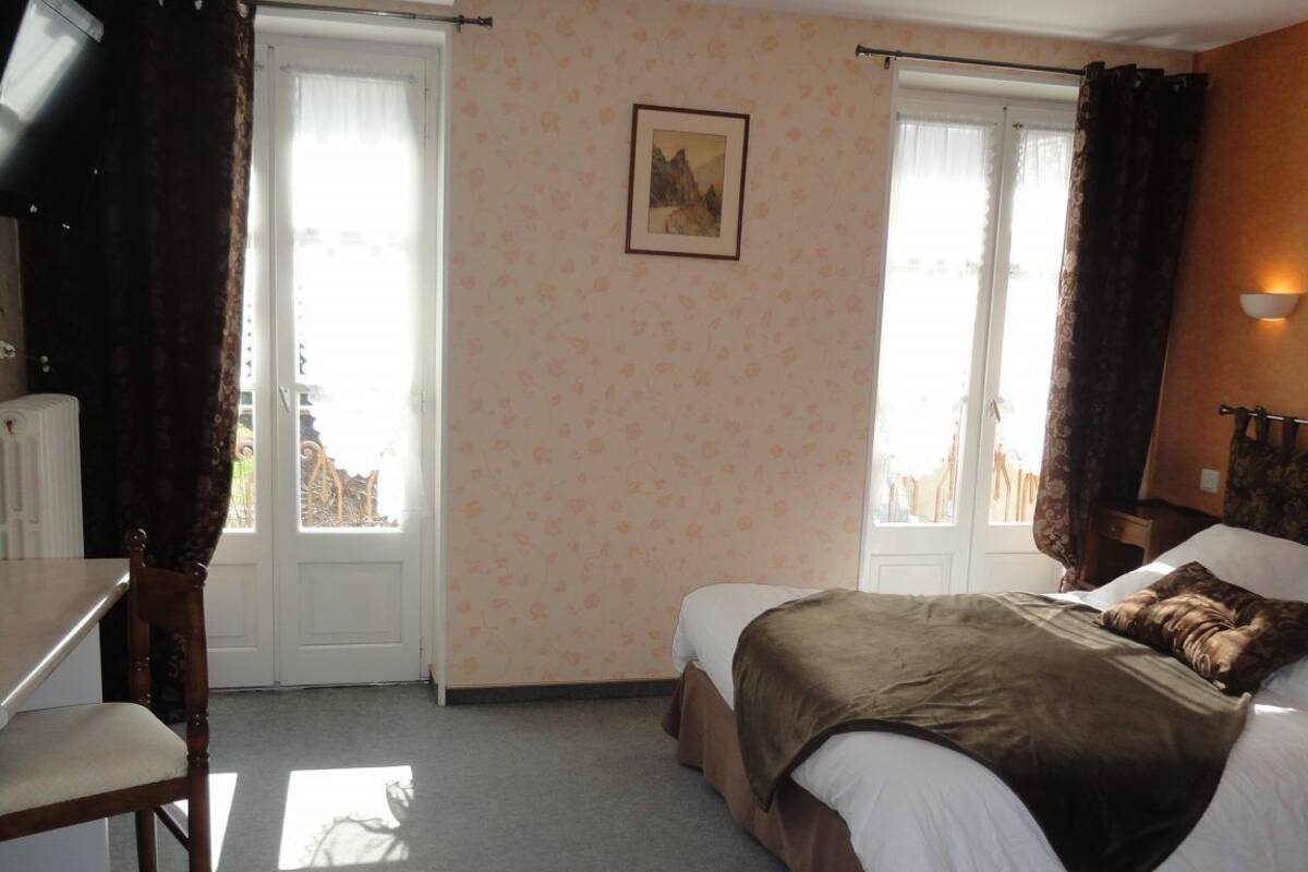 A hotel room with a bed, a desk, and a chair. There are two windows with curtains, a door, and a wall-mounted picture.