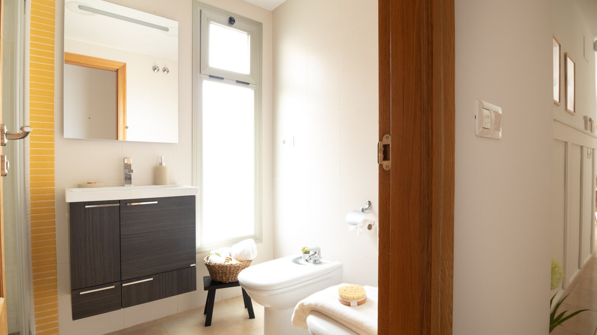 Une salle de bain moderne avec un meuble-lavabo en bois foncé, un lavabo blanc et des toilettes. Une fenêtre laisse entrer la lumière naturelle, et on aperçoit une porte en bois.