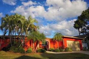 A red house with a garage, surrounded by palm trees and a clear sky.