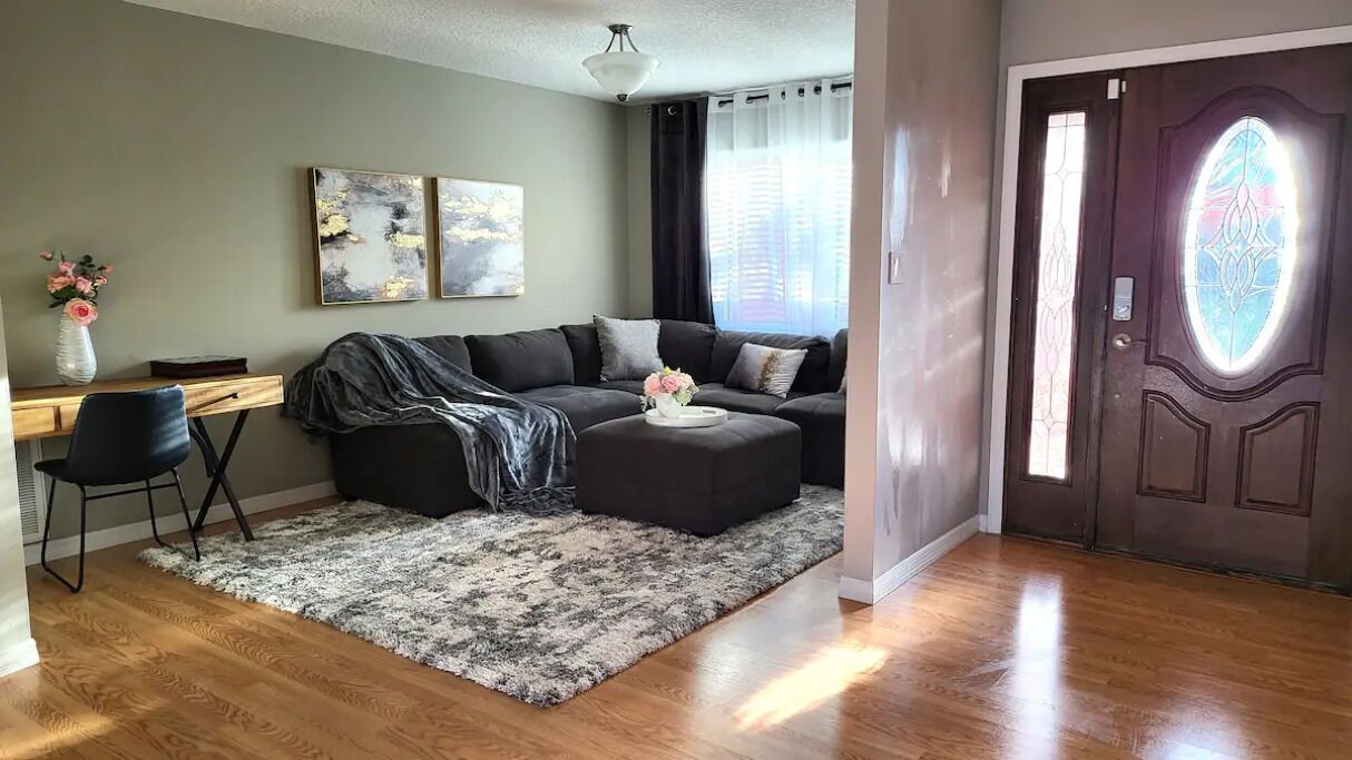 Una sala de estar con un sofá de color oscuro, una mesa de centro y una alfombra. Hay un escritorio con una silla y un jarrón de flores. El cuarto tiene piso de madera y una puerta con un vitral.