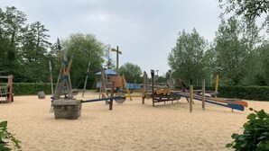 Une aire de jeux dotée de divers équipements, notamment des balançoires et des structures d’escalade, aménagée sur un sol sablonneux avec des arbres en arrière-plan.