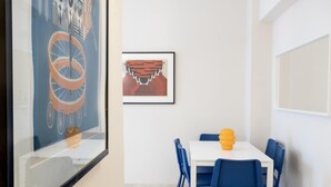 Ein Essbereich mit einem weißen Tisch und blauen Stühlen, einem gerahmten Kunstwerk an der Wand und einem Fenster mit Blick auf ein Gebäude.