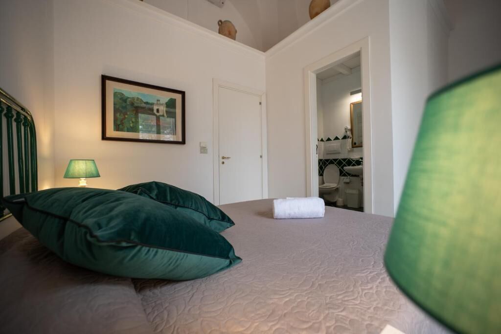 Et soveværelse med seng, puder, en lampe, et billede i ramme og en dør til et andet rum.