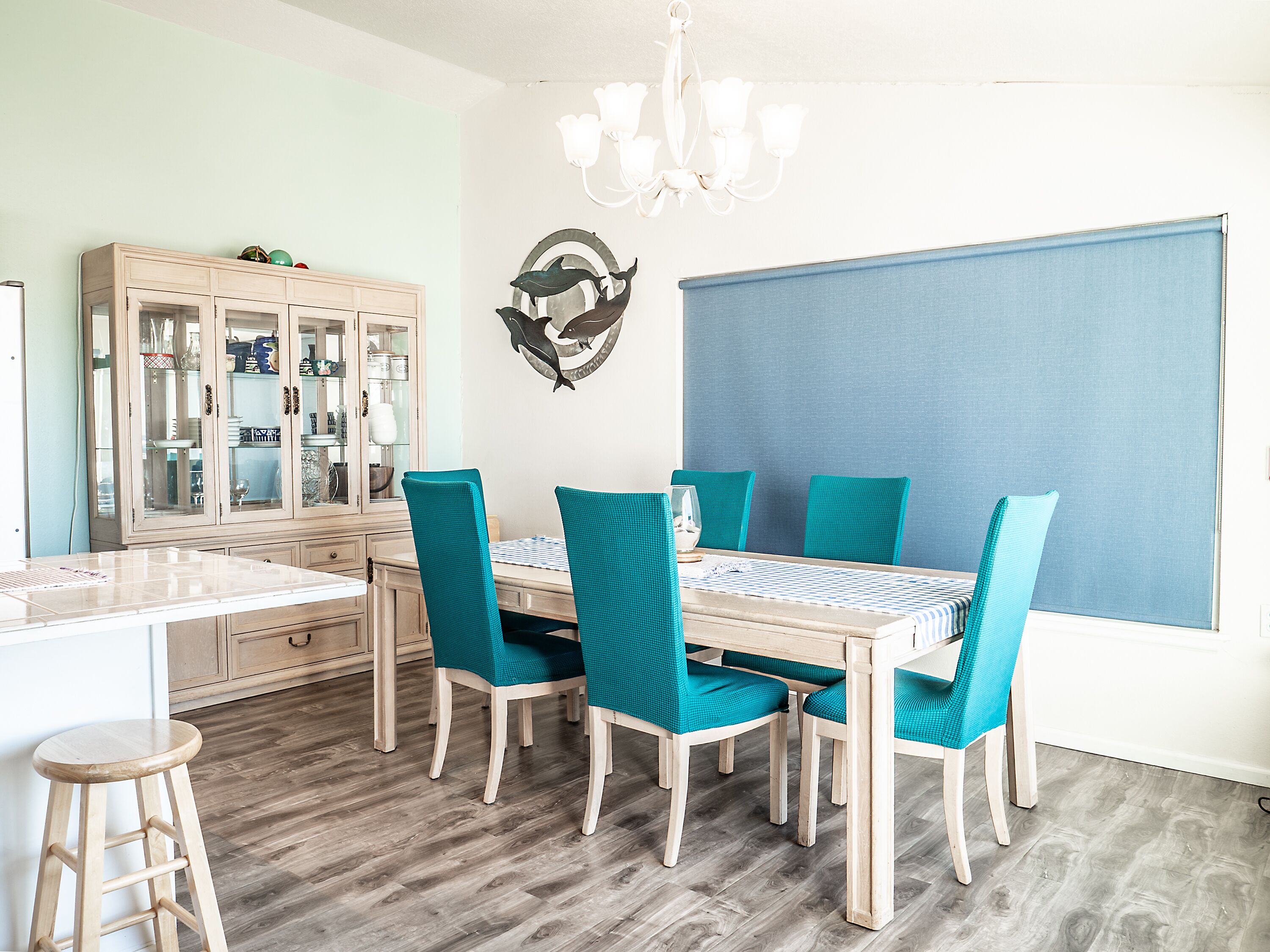 A dining area with a wooden table, teal upholstered chairs, a glass cabinet, and a wall-mounted dolphin decal.
