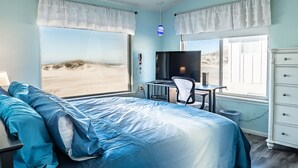 Ein Schlafzimmer mit einem großen Bett, einem Schreibtisch mit Computer, einem Fernseher und Blick auf den Strand.