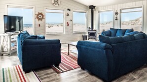 Ein Wohnzimmer am Meer mit Fernseher, blauen Sofas, Kamin und großen Fenstern mit Blick auf den Strand.