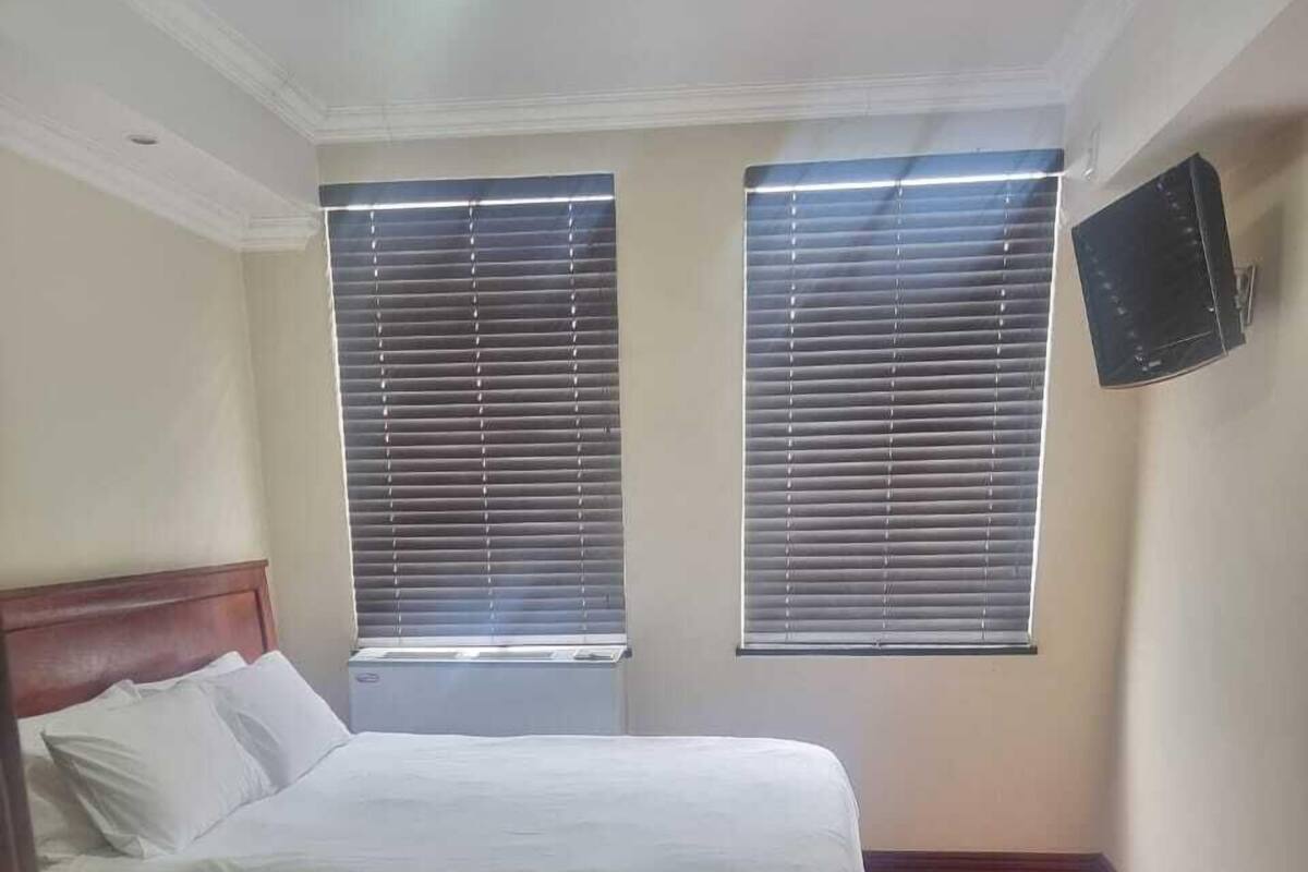 A bedroom with a bed, two windows with blinds, a red carpet, and a mounted TV on the wall.