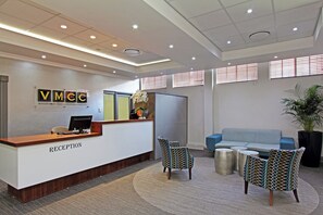 A reception area with a reception desk labeled "RECEPTION," a blue sofa, patterned chairs, and a potted plant. - VMCC (Johannesburg)