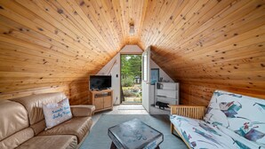 Un acogedor dormitorio en el ático con techo inclinado de madera, un sofá, una mesa de centro y un televisor sobre un mueble de madera.