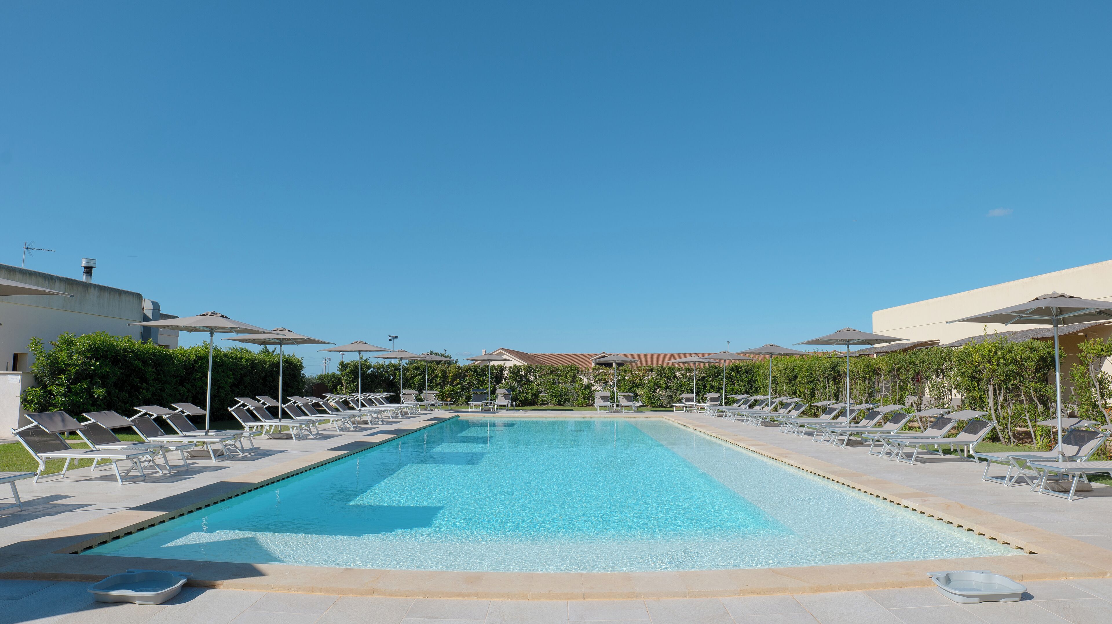 A rectangular swimming pool with clear blue water, surrounded by lounge chairs and umbrellas, and a backdrop of greenery and buildings.