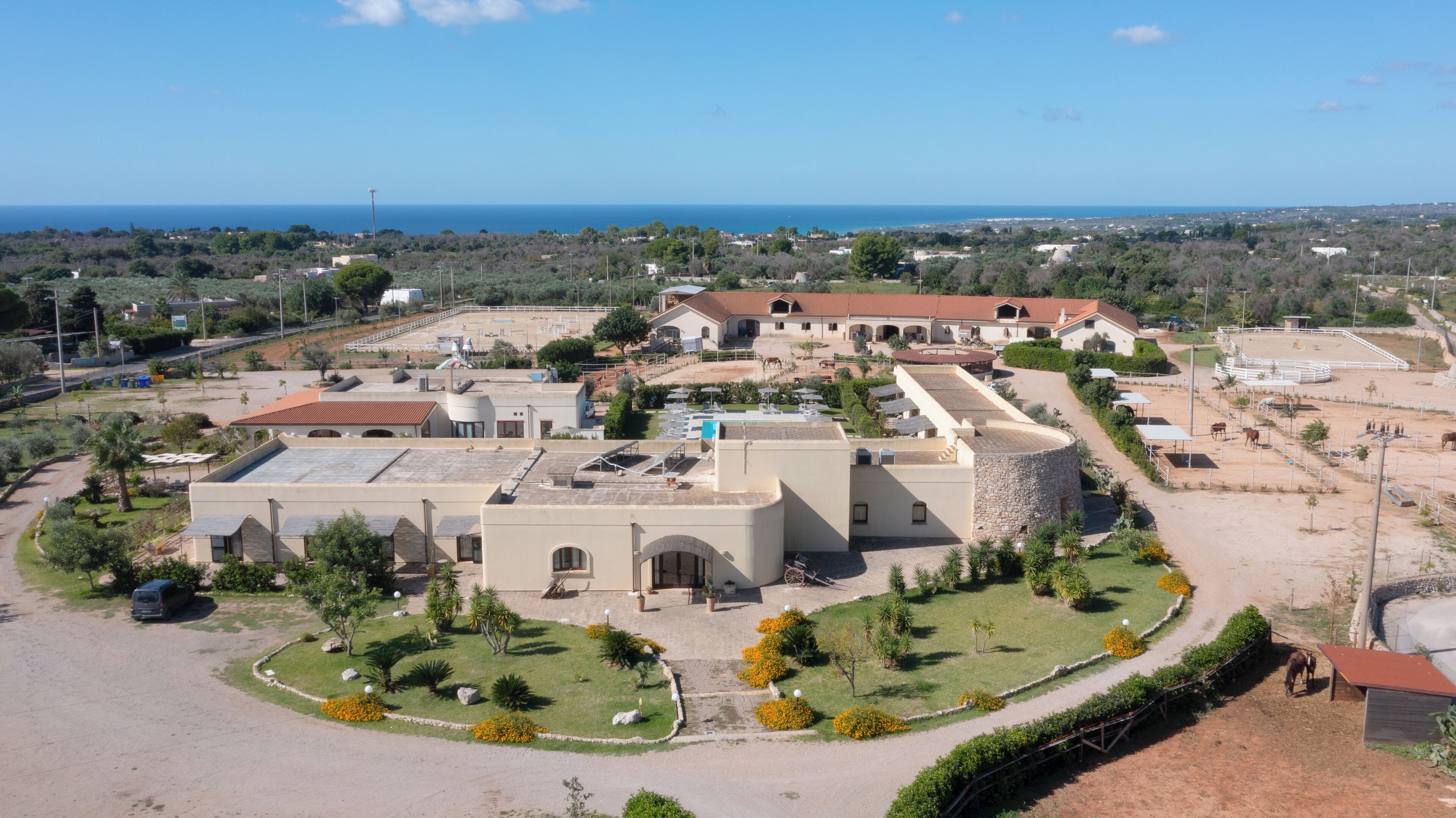 Une vue aérienne d’une propriété comprenant plusieurs bâtiments, dont une grande structure centrale dotée d’un toit plat, entourée de jardins aménagés et d’un espace clos où se trouvent des chevaux.