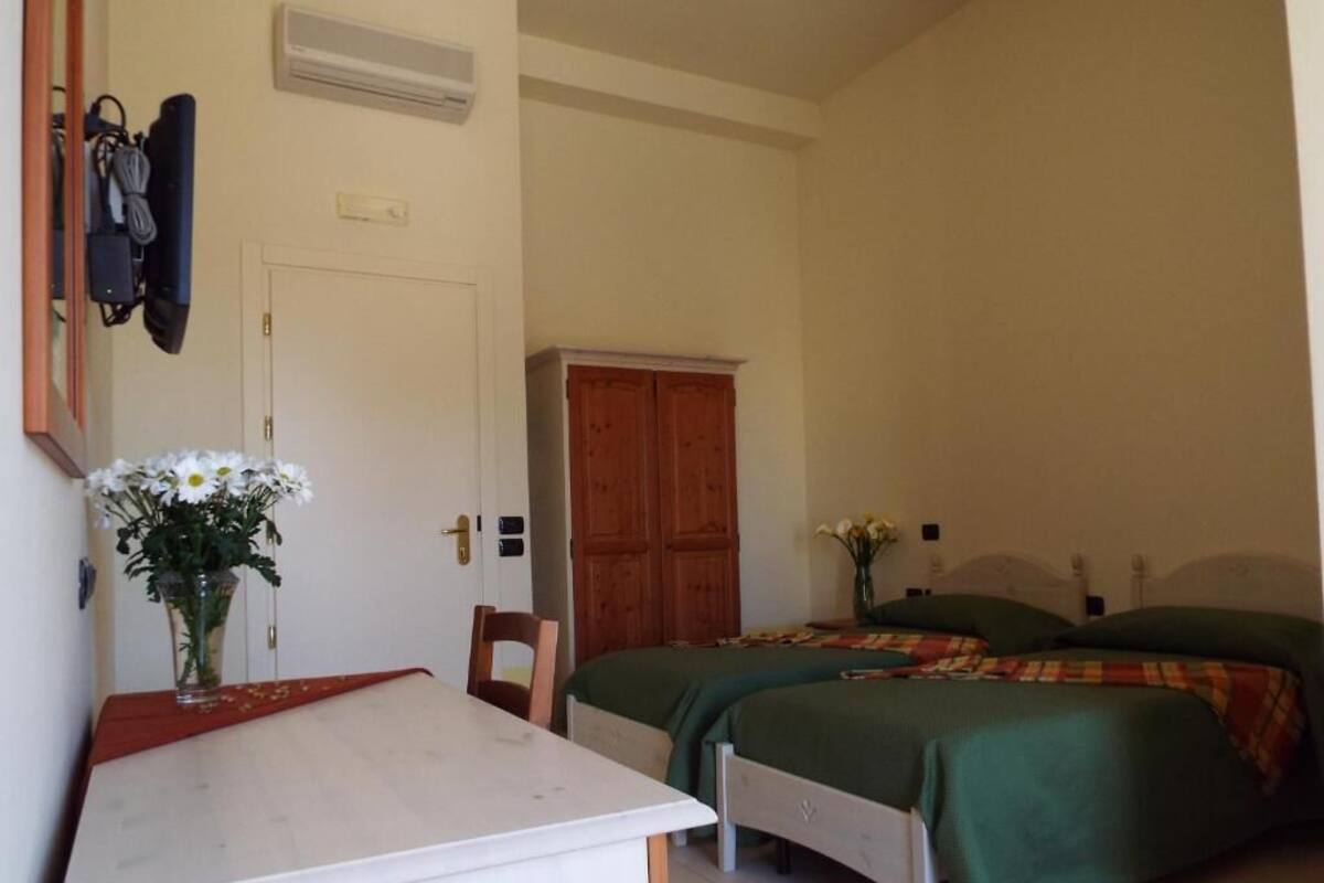 A bedroom with two beds, a wooden wardrobe, a desk with a chair, and a vase with flowers.