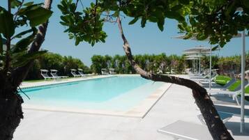 Une piscine entourée d’arbres, avec des chaises longues et des parasols, sous un ciel dégagé.