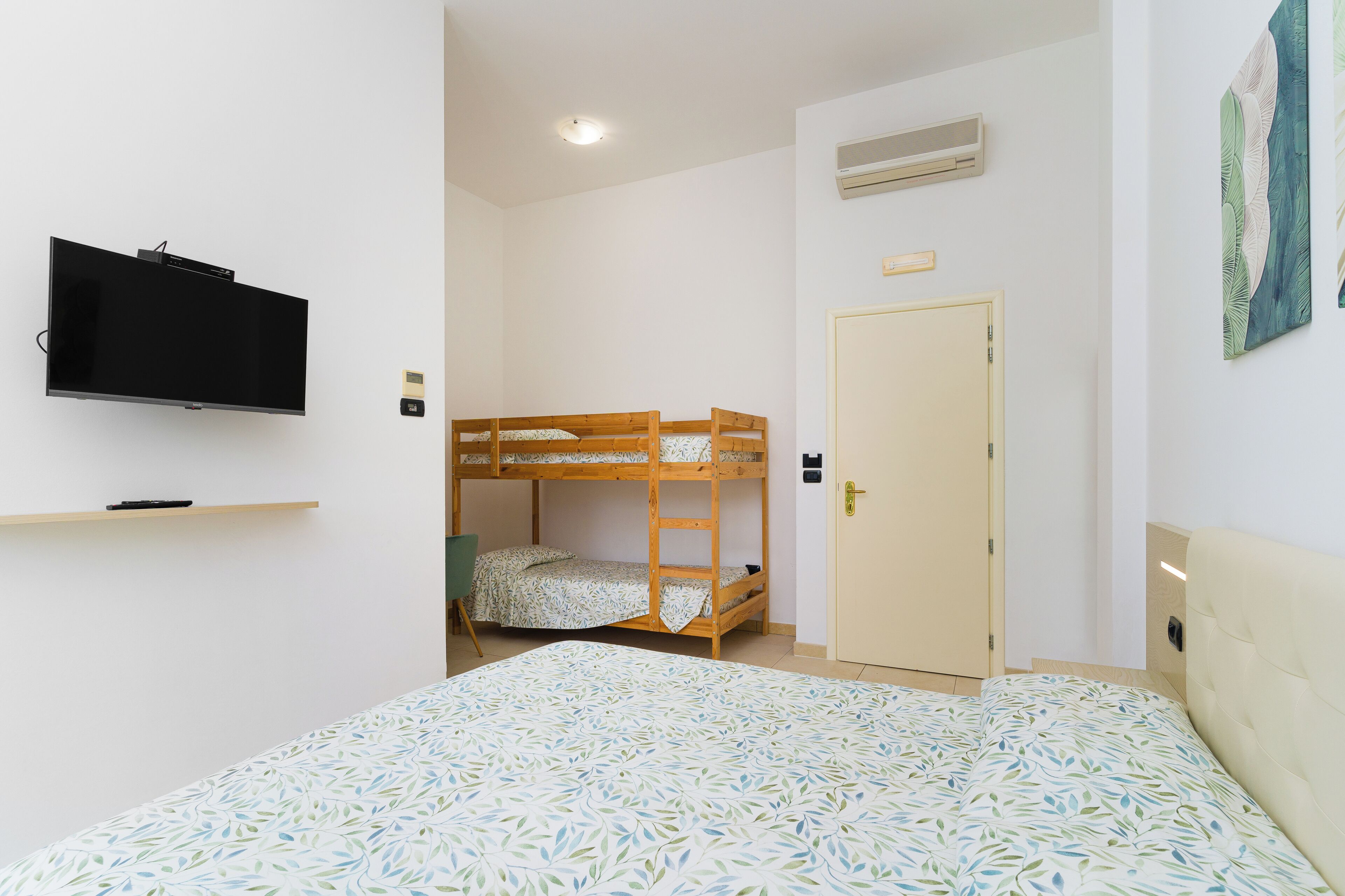 Quarto com beliche, televisão na parede, uma porta e cama com colcha estampada.