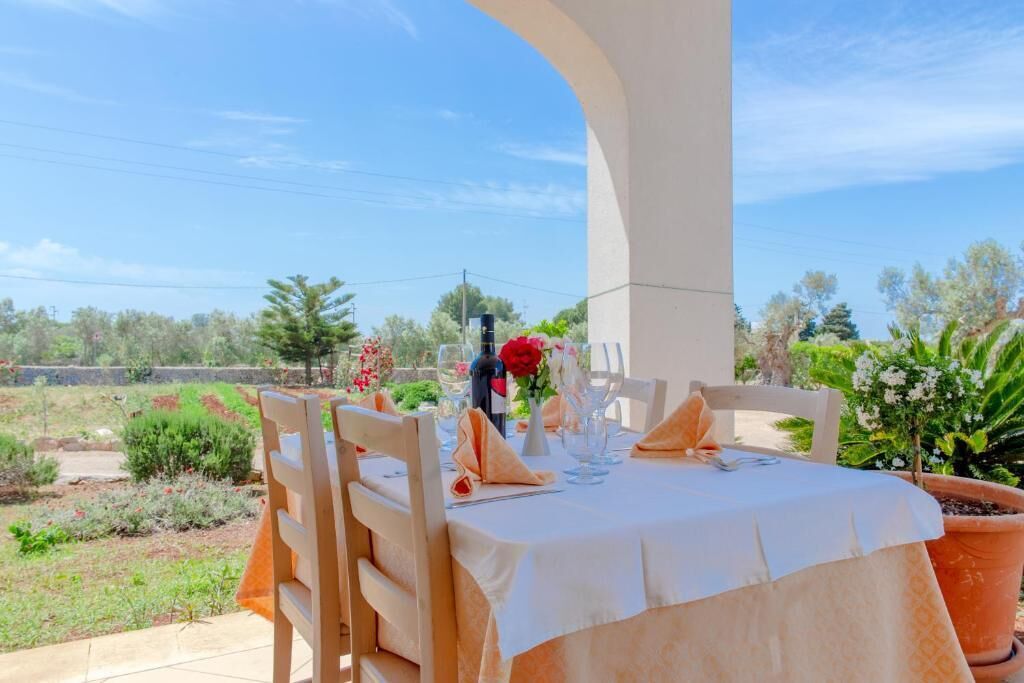 An outdoor dining area with a table set for two, surrounded by chairs, with a view of a garden and trees in the background.