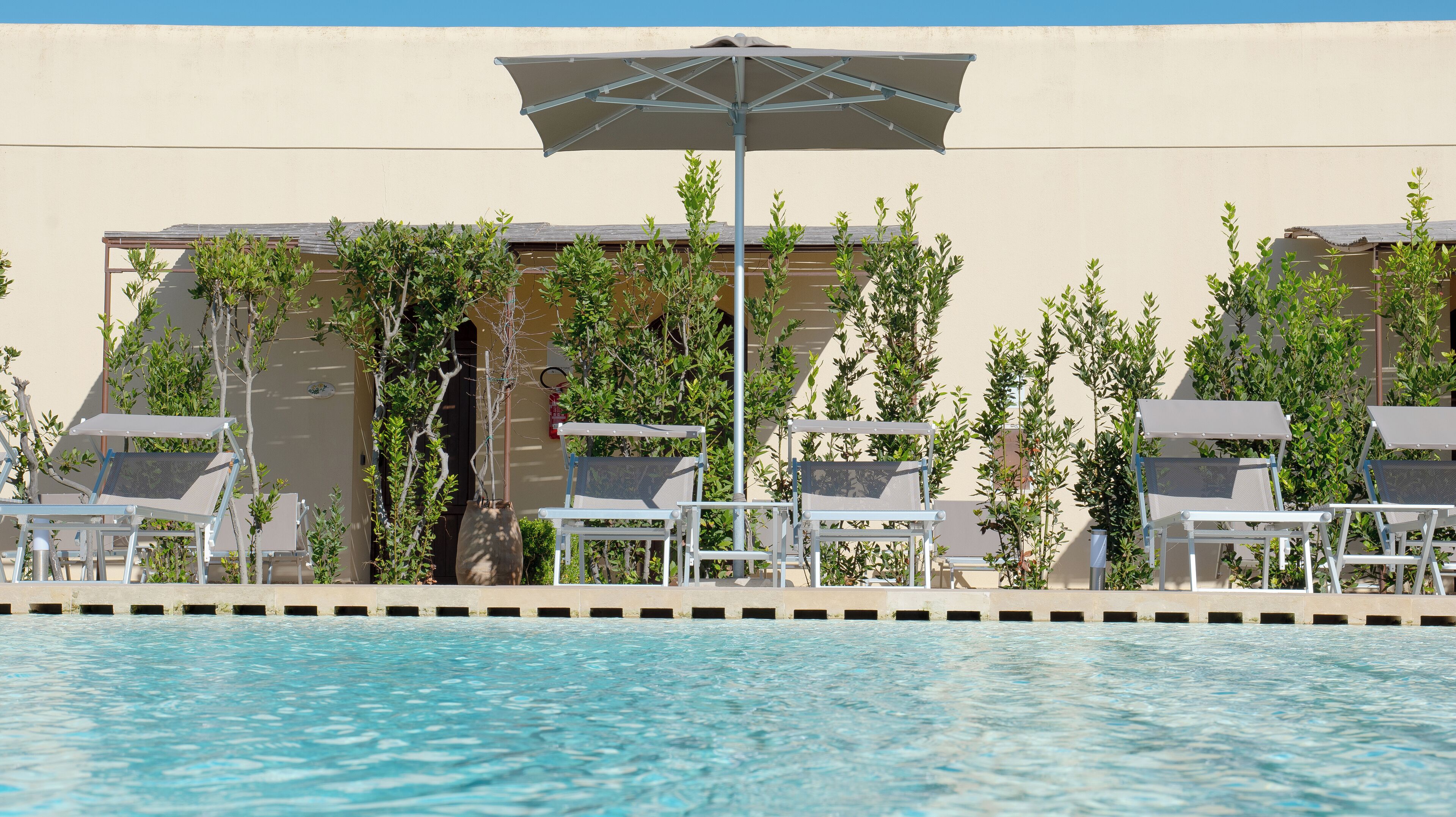 A poolside area with lounge chairs and umbrellas, surrounded by greenery and a building facade.
