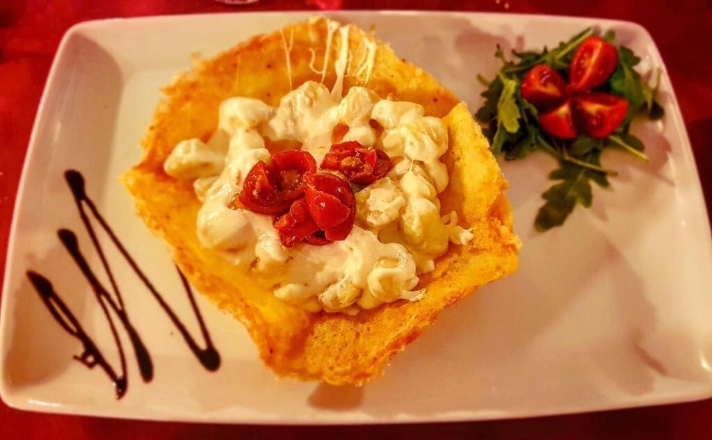 Een vierkant bord met een dessert, afgetopt met slagroom en rode vruchten, vergezeld van een kleine salade en chocoladesiroop.
