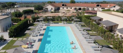 A large rectangular swimming pool with lounge chairs and umbrellas around it, surrounded by buildings and greenery.