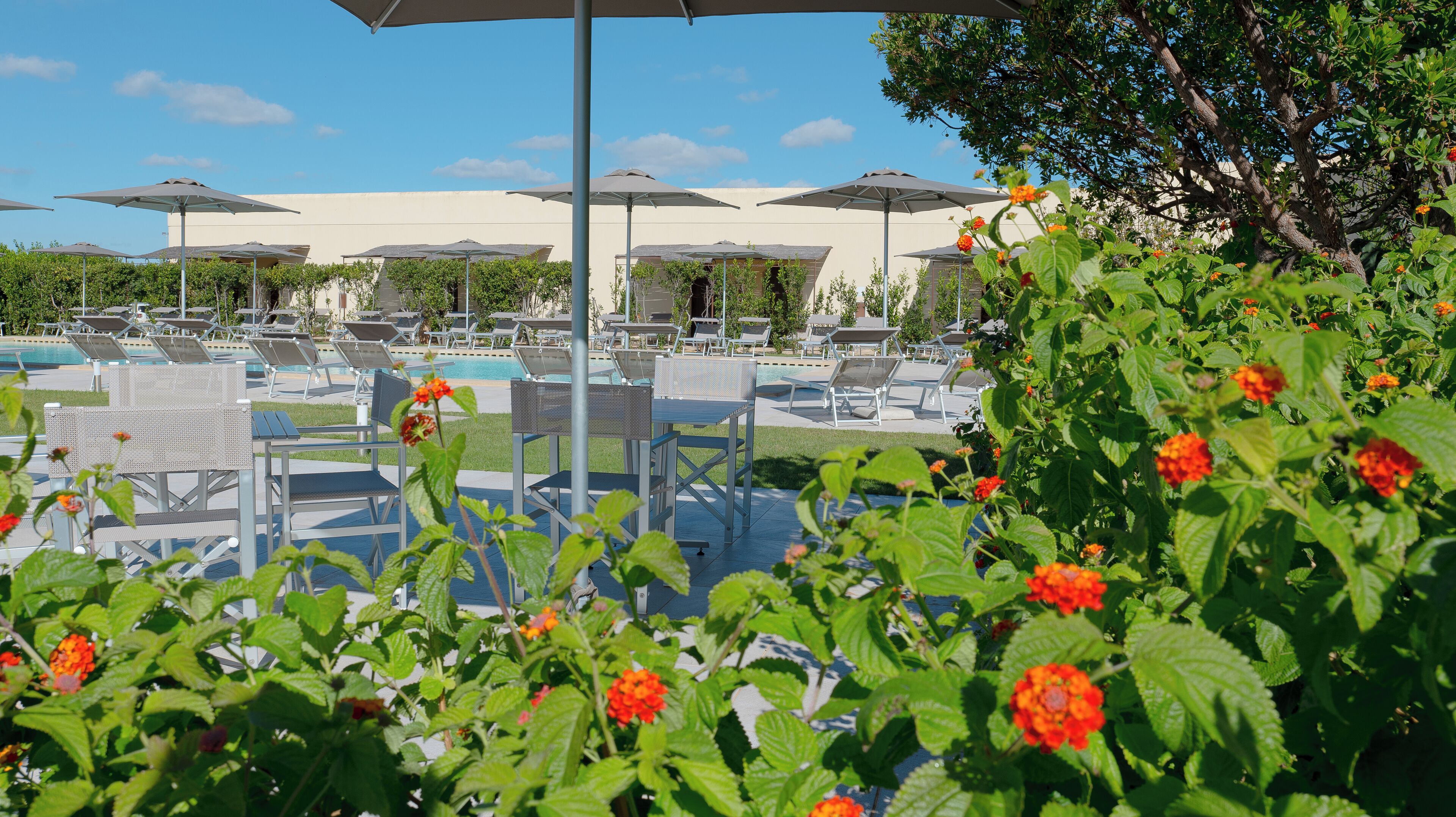 A poolside area with lounge chairs and umbrellas, surrounded by greenery and flowers.