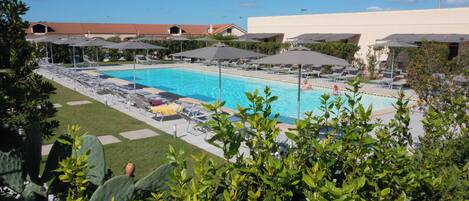 A pool area with lounge chairs and umbrellas, surrounded by greenery and buildings in the background.
