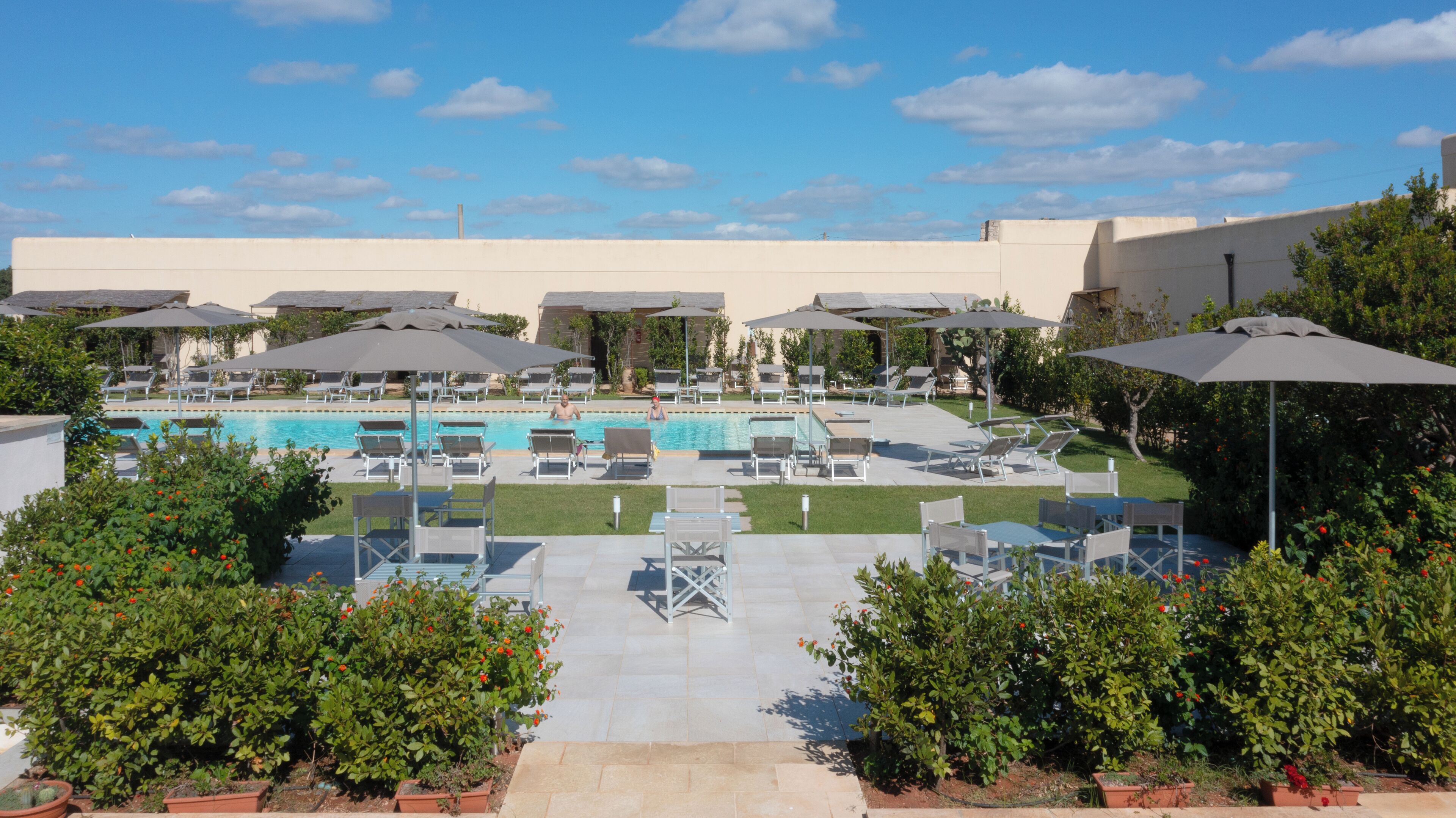 A pool area with lounge chairs and umbrellas, surrounded by greenery and a building in the background.