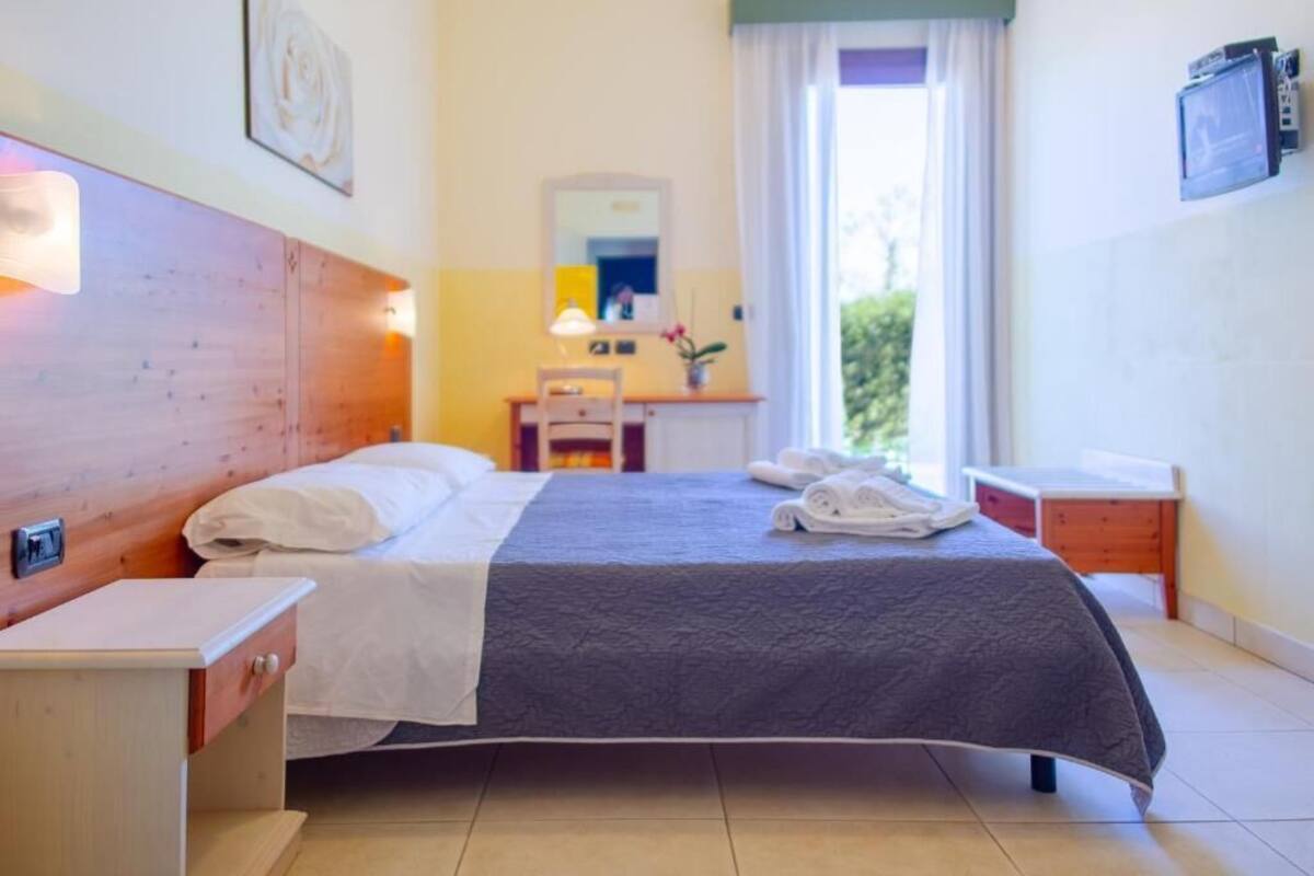 A hotel room with a wooden headboard, a bed with white linens, a bedside table, a desk with a chair, a mirror, and a window with curtains.