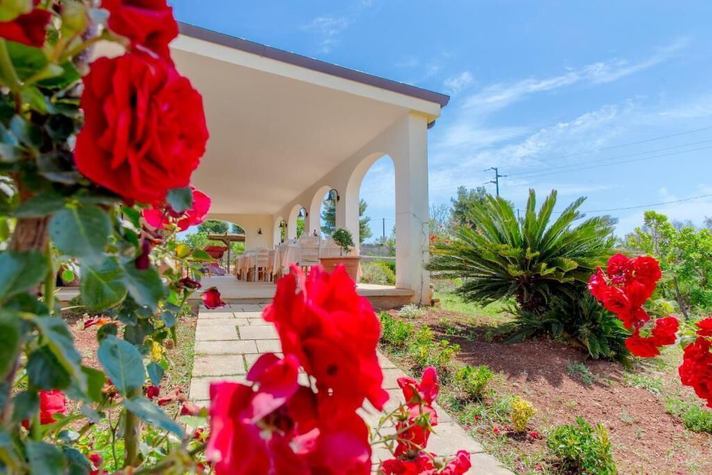 Un espacio de estar al aire libre cubierto con una estructura blanca y arcos, rodeado de flores rojas y follaje.