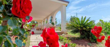 Un espacio de estar al aire libre cubierto con una estructura blanca y arcos, rodeado de flores rojas y follaje.