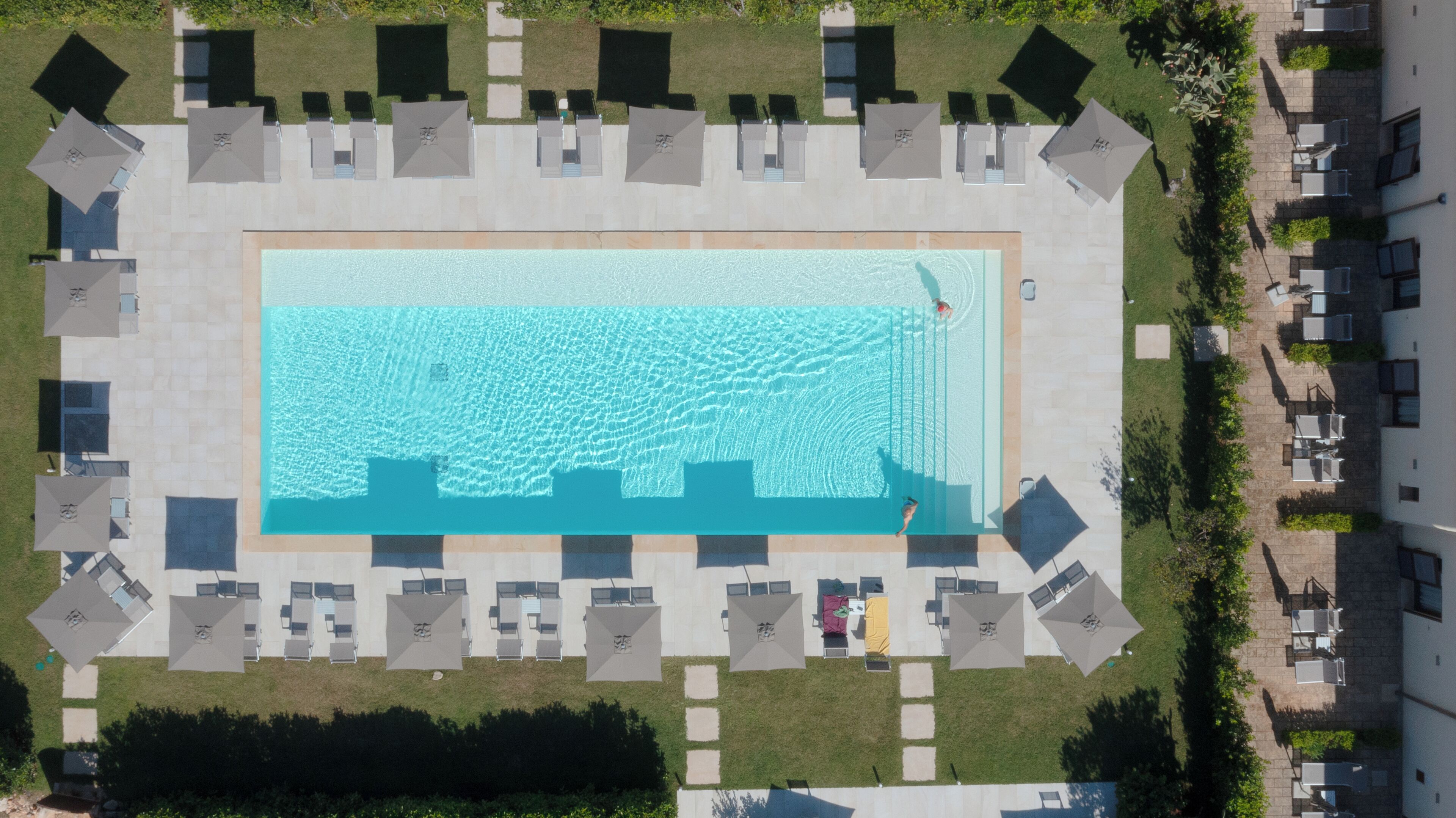 An aerial view of a rectangular swimming pool surrounded by lounge chairs and umbrellas, with a paved walkway and greenery.