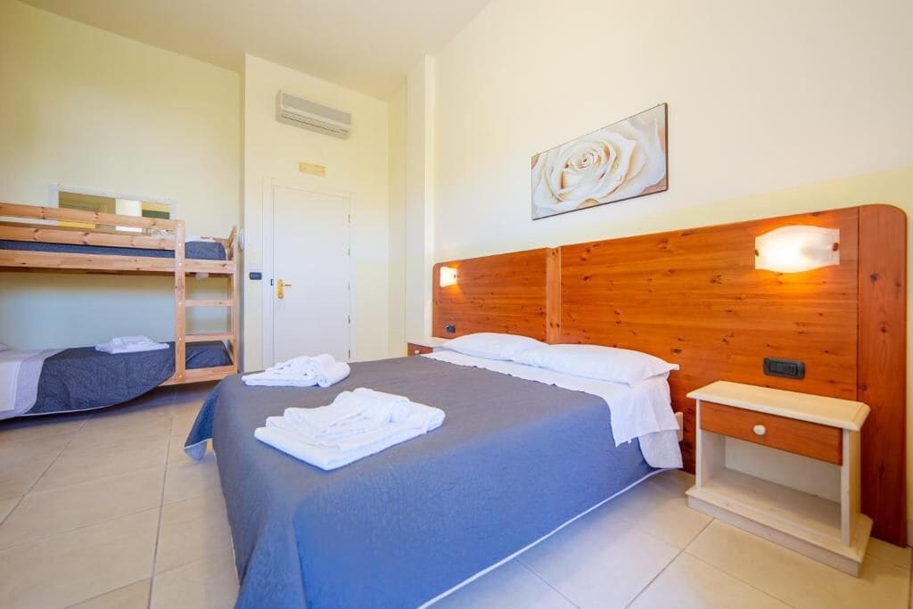 A room with a large bed, a smaller bunk bed, a bedside table, and a wall-mounted light fixture.