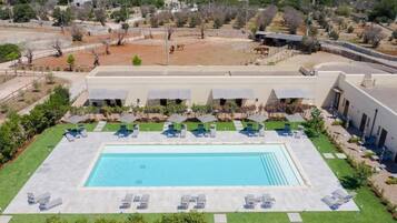 A large rectangular swimming pool with lounge chairs and umbrellas, surrounded by a building with multiple rooms and a fenced area with animals.