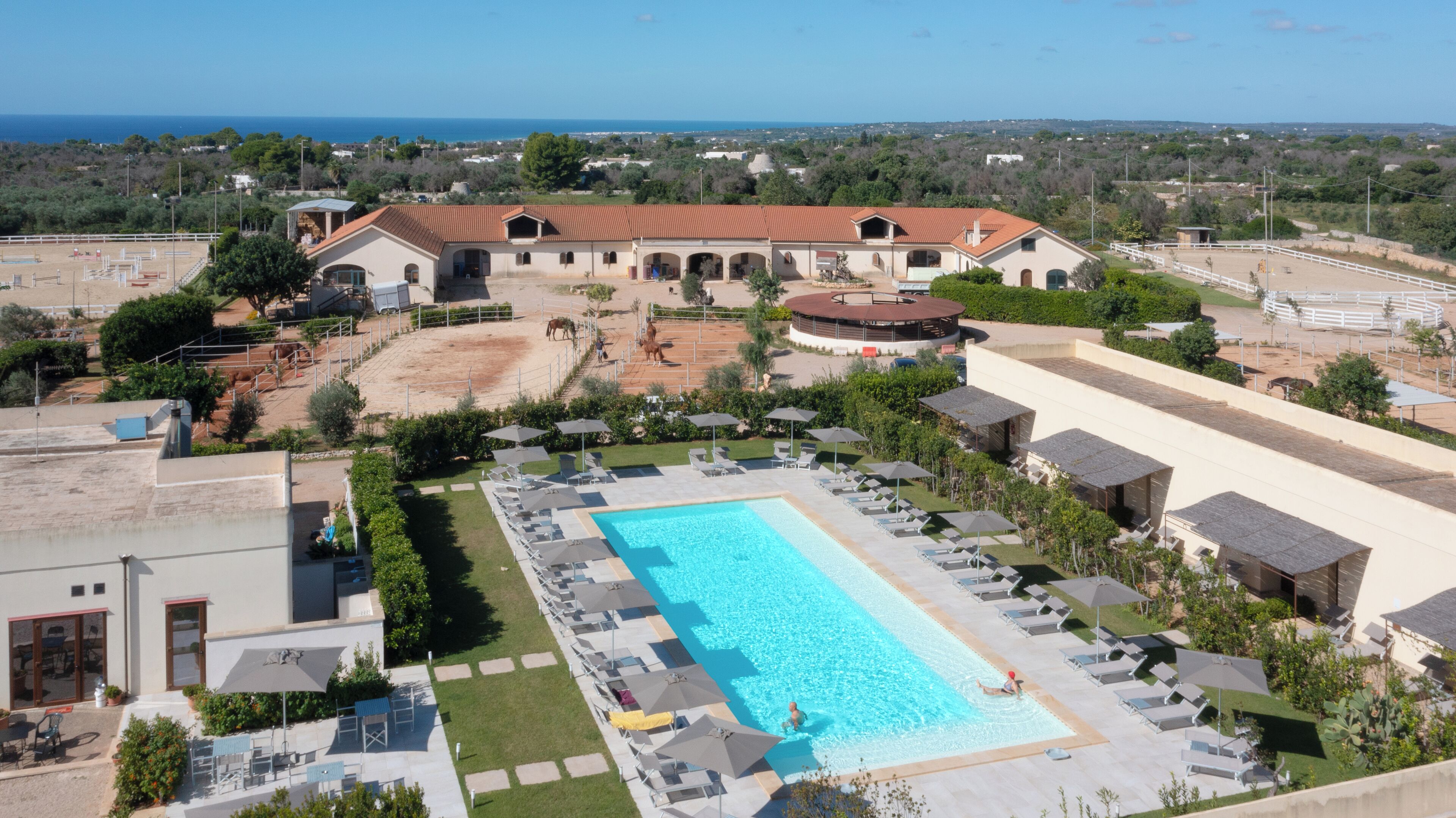 A large swimming pool with sun loungers and umbrellas, surrounded by a landscaped area with a view of the sea.