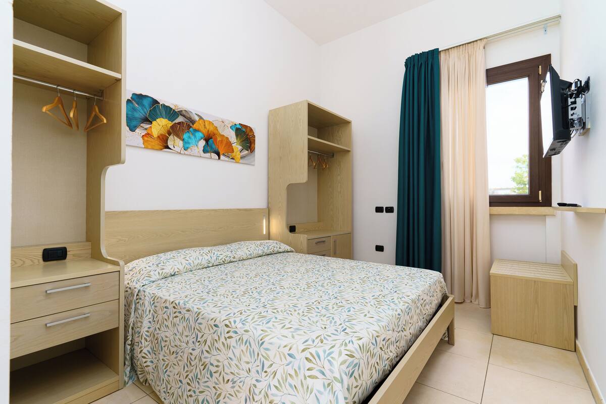 A bedroom with a bed, a wooden nightstand, a dresser, a mirror, and a wall-mounted TV.