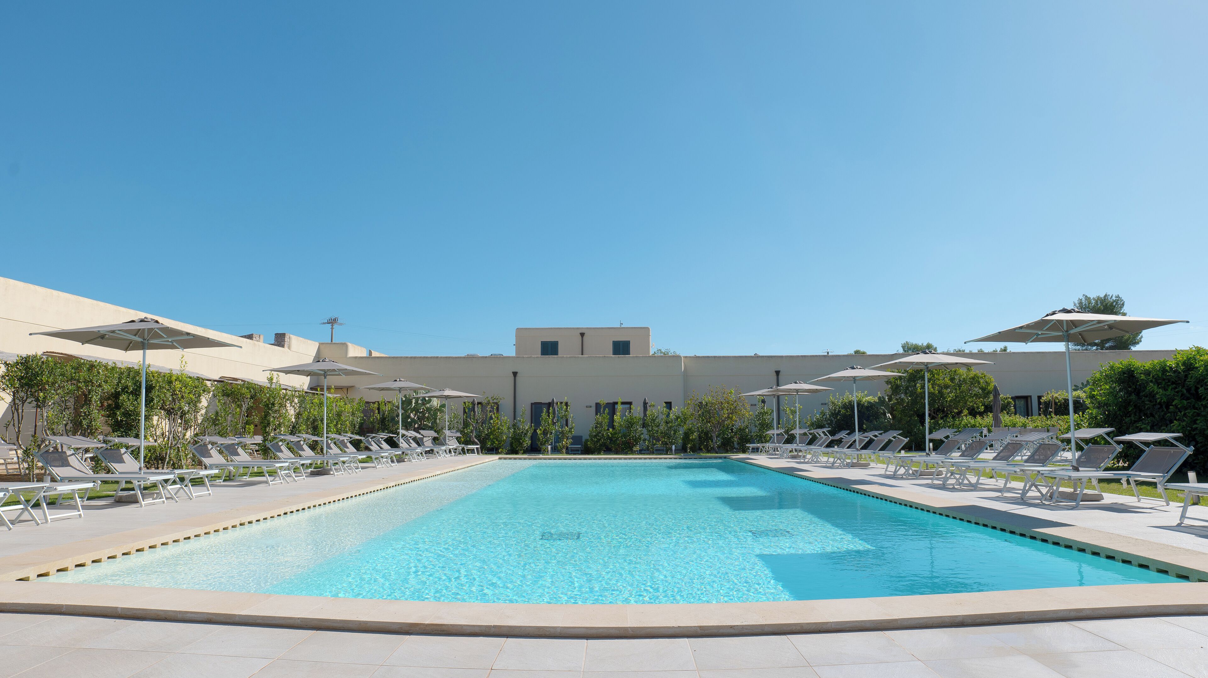 Una piscina con sillas de descanso y sombrillas, rodeada por un edificio de techo plano y ventanas.