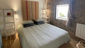 Een slaapkamer met een houten plafond, een stenen muur, een bed met kussens, een nachtkastje en een raam met gordijnen.