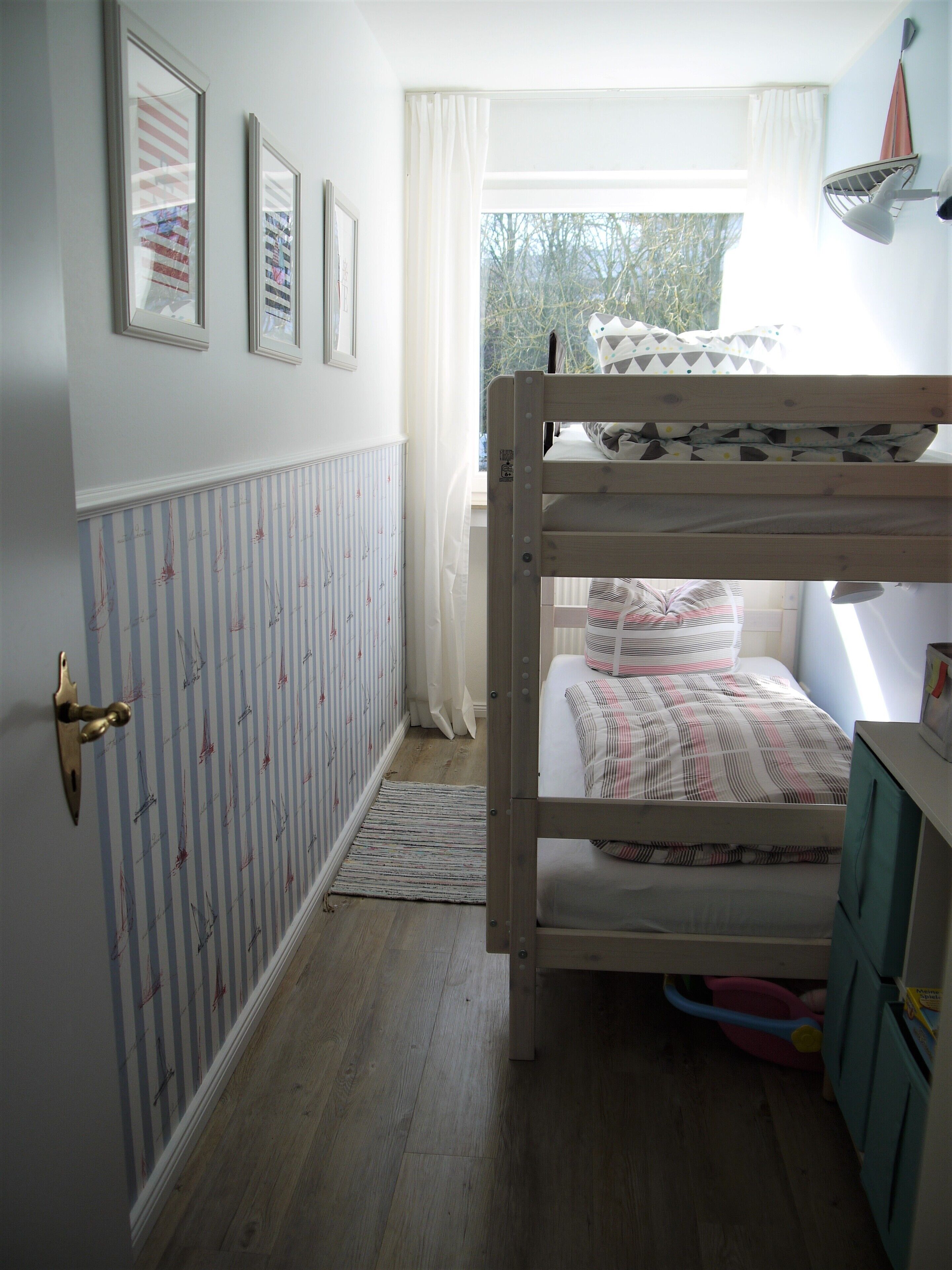 Un dormitorio infantil con literas, un radiador, una ventana con cortinas y una puerta con picaporte de latón.