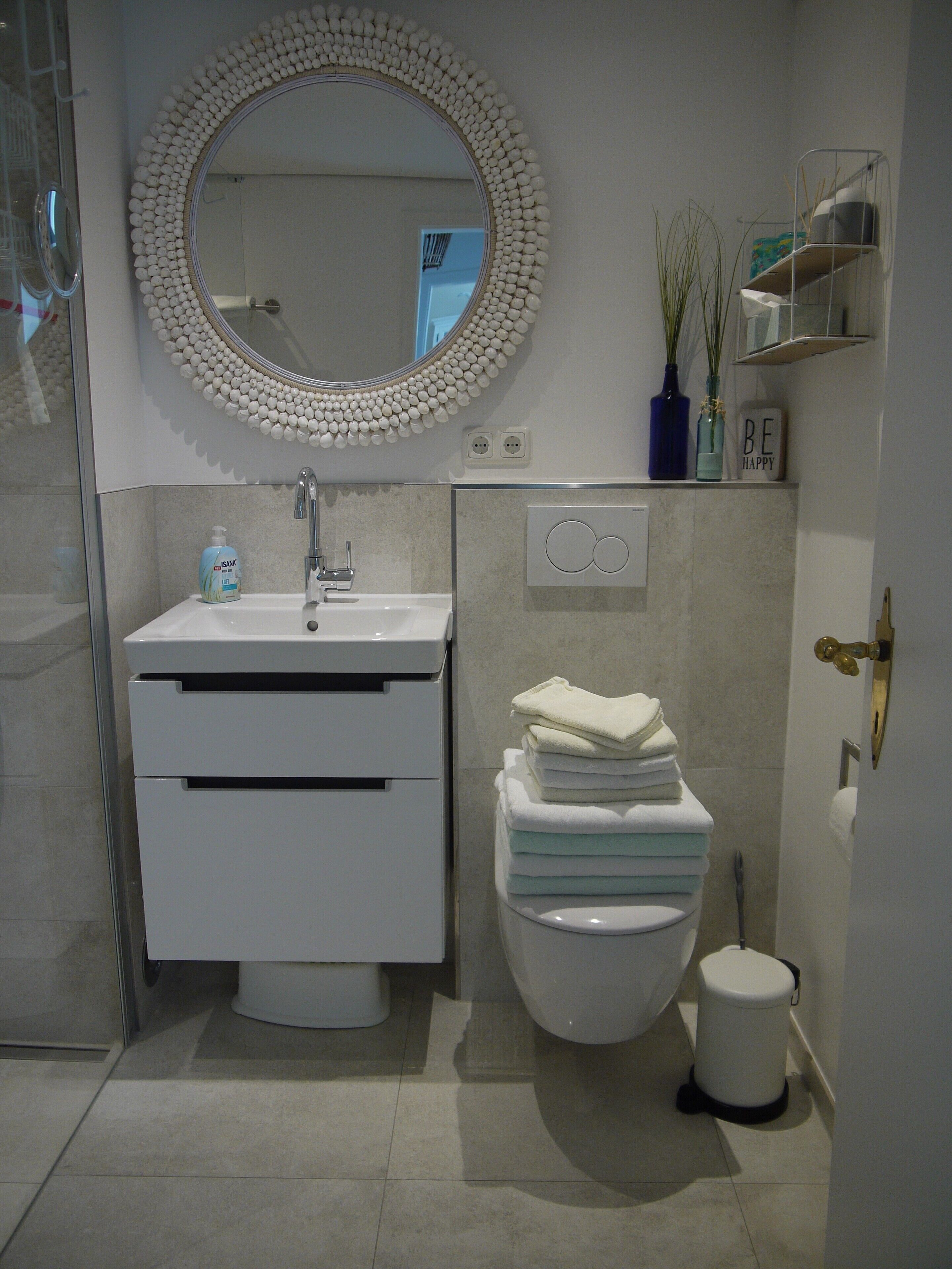 Een moderne badkamer met een ronde spiegel, een witte wasbak en toilet. Handdoeken liggen op het toilet gestapeld, er is een prullenbak en een klein schapje met decoratieve voorwerpen.
