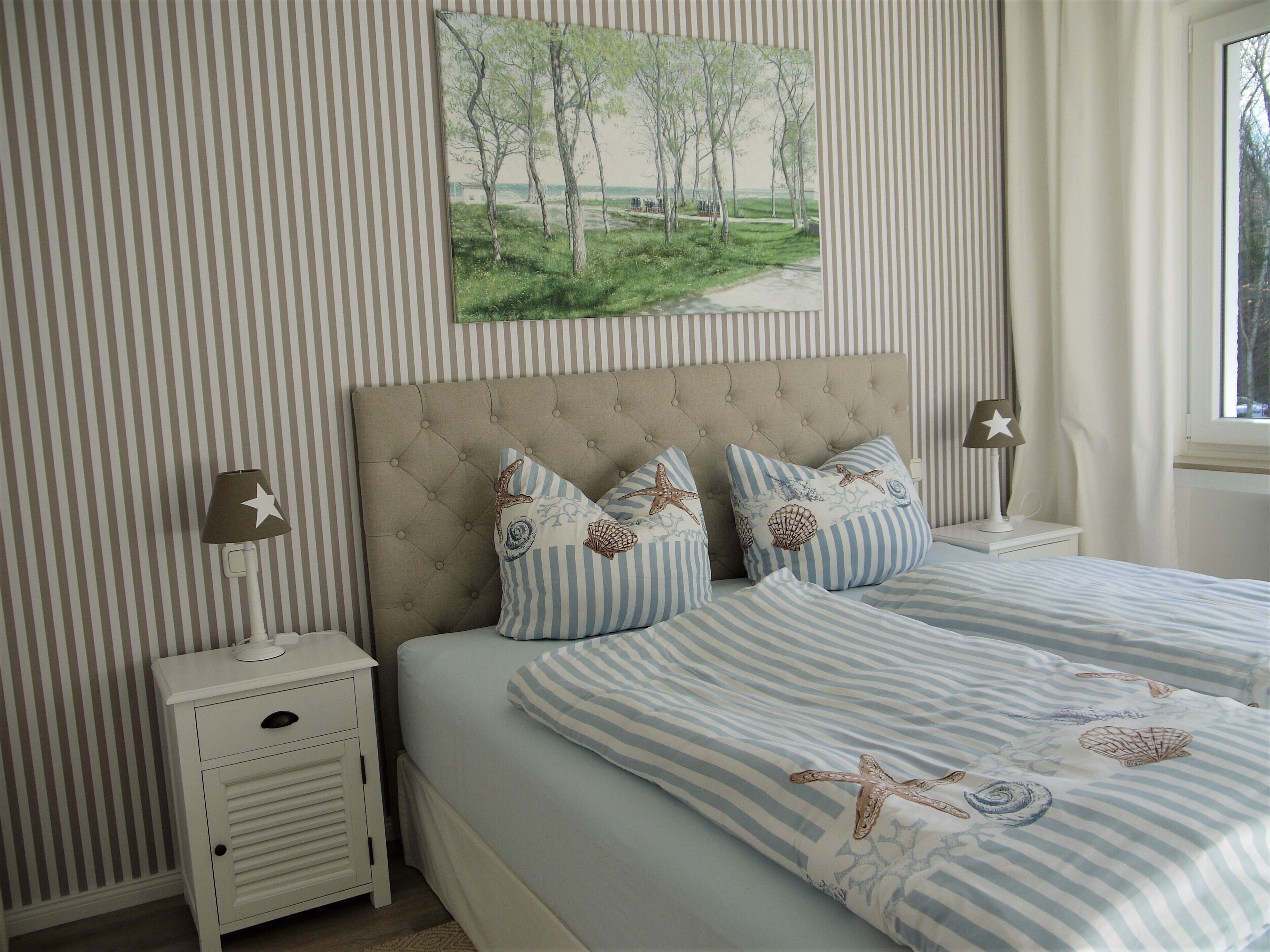 Une chambre avec un papier peint à rayures, une tête de lit capitonnée et un tableau représentant des arbres.
