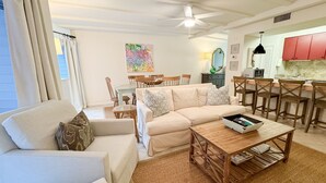 A living room with a sofa, coffee table, and dining area with chairs. - The Olive Shell- condo steps to the beach (St Simons Island)