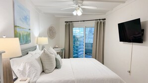 A hotel room with a bed, a desk, a chair, a TV, a ceiling fan, and a window with curtains. - The Olive Shell- condo steps to the beach (St Simons Island)