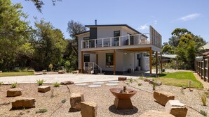Une maison moderne à deux étages, dotée d’un balcon et entourée d’un jardin agrémenté de rochers et de plantes.