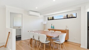 Une cuisine moderne avec des meubles blancs, une table à manger en bois et des chaises blanches, et une vue sur l’extérieur par une fenêtre.