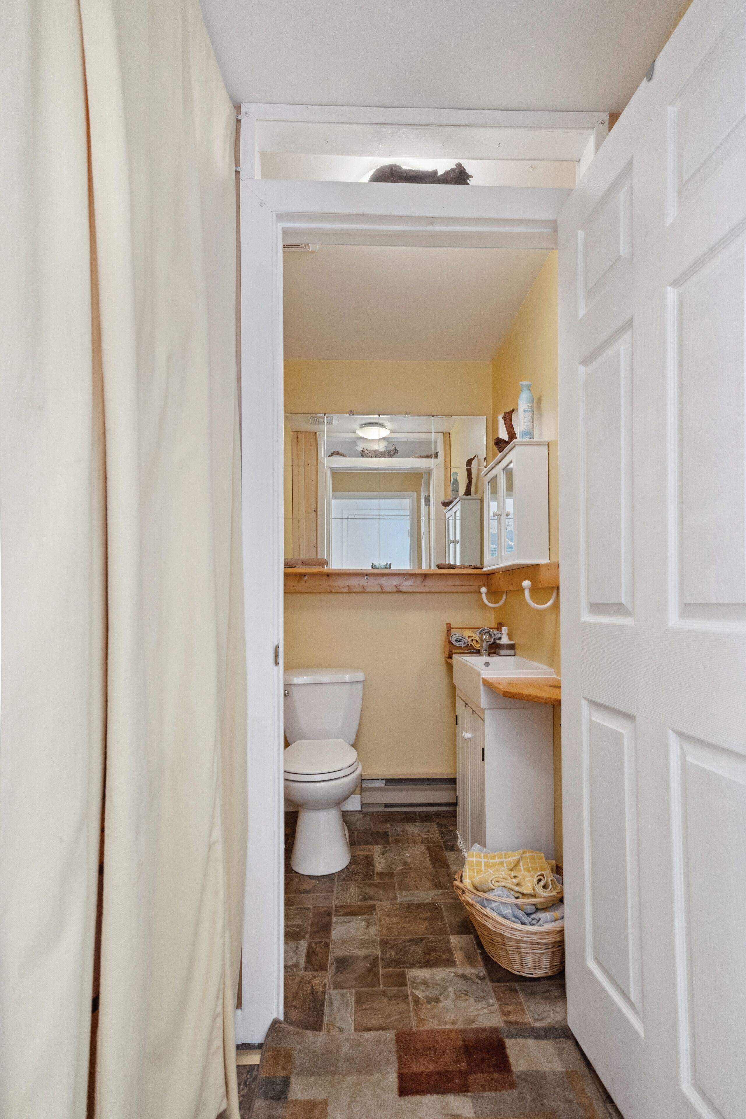Ein schmaler Flur, der zu einem Badezimmer mit Toilette, Waschbecken und einem Wäschekorb führt.